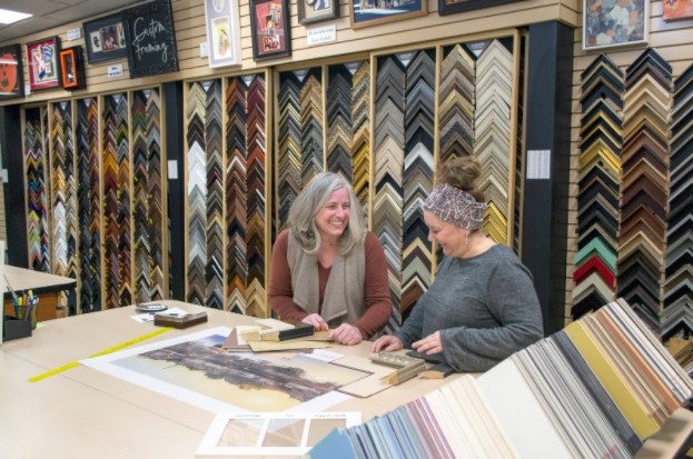 Two women laughing and conversing inside the framing shop, surrounded by walls filled with various frame designs